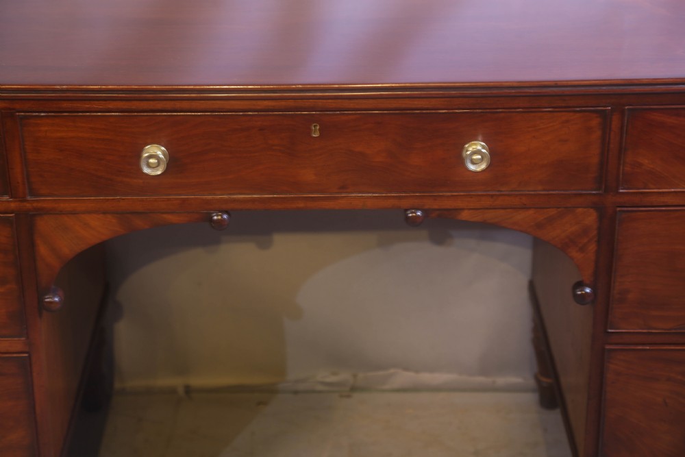 A 19th-century mahogany kneehole desk featuring multiple drawers with brass knobs, a central arched apron, and turned legs. View VI