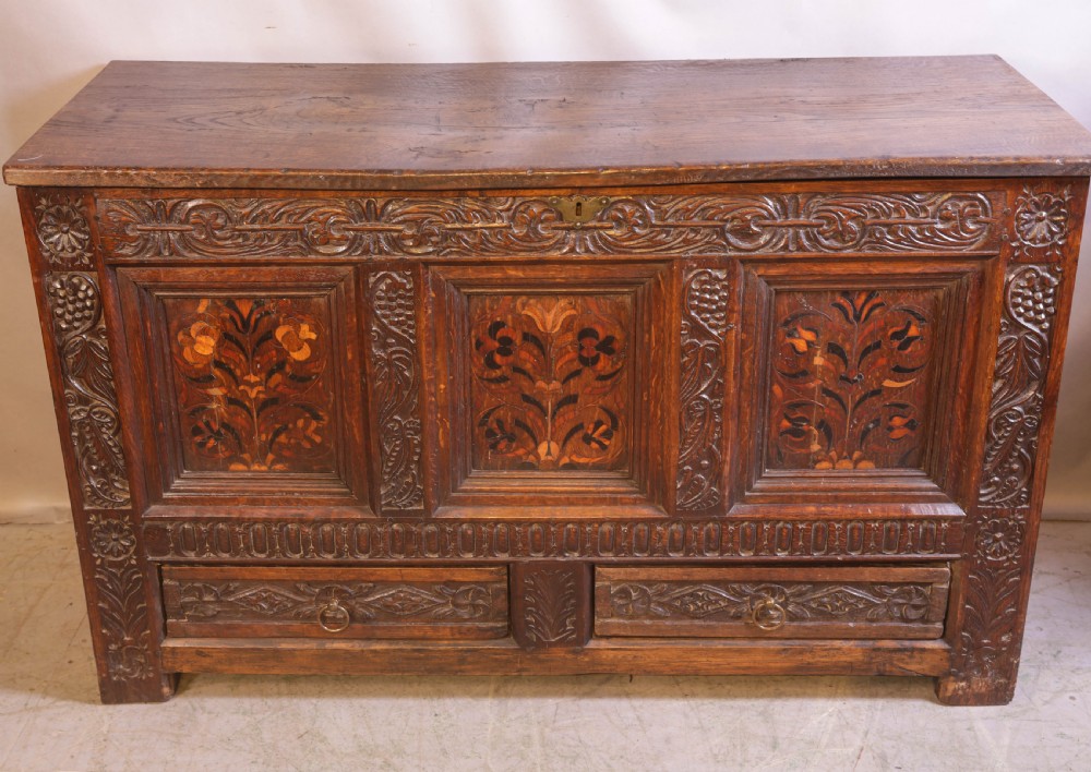 An antique carved oak coffer featuring three floral inlaid panels, carved frieze work, and two lower drawers with brass handles.