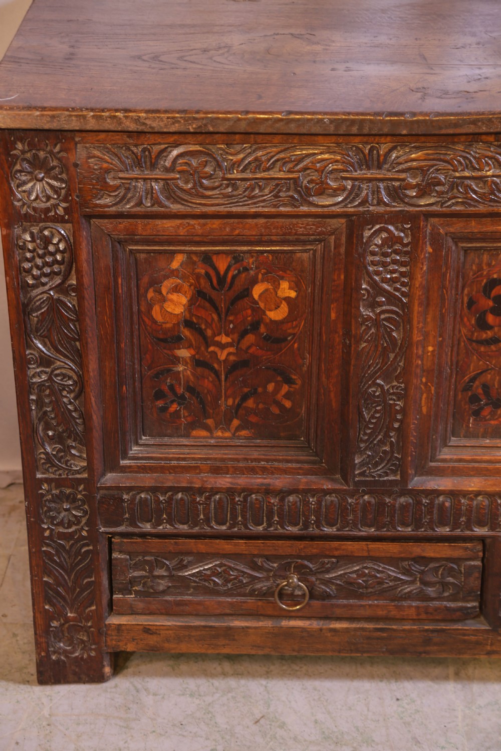 An antique carved oak coffer featuring three floral inlaid panels, carved frieze work, and two lower drawers with brass handles. View V