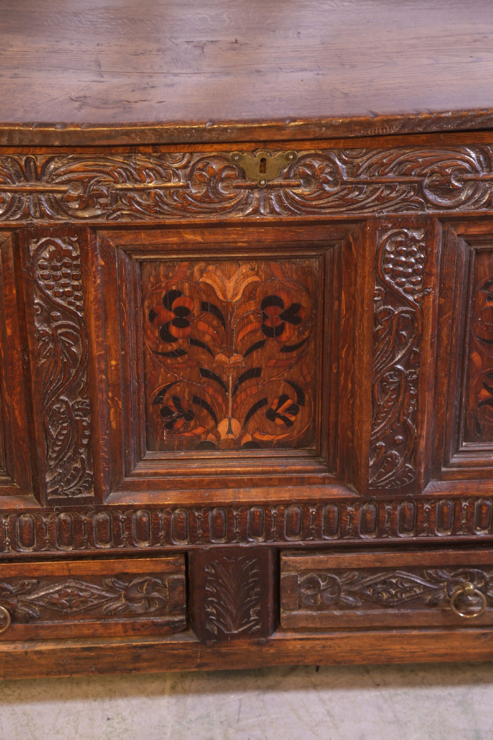 An antique carved oak coffer featuring three floral inlaid panels, carved frieze work, and two lower drawers with brass handles. View VII