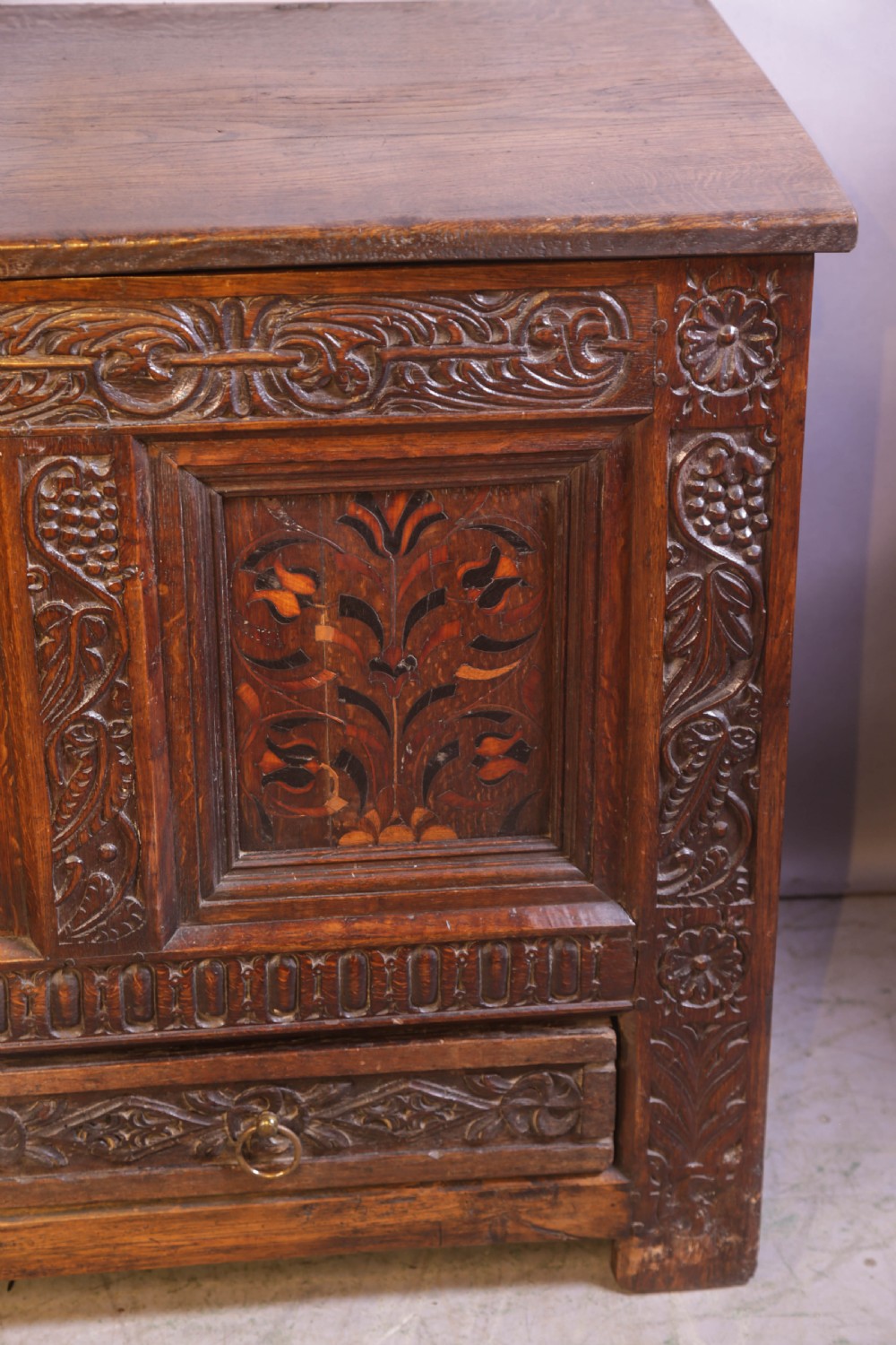 An antique carved oak coffer featuring three floral inlaid panels, carved frieze work, and two lower drawers with brass handles. View IX
