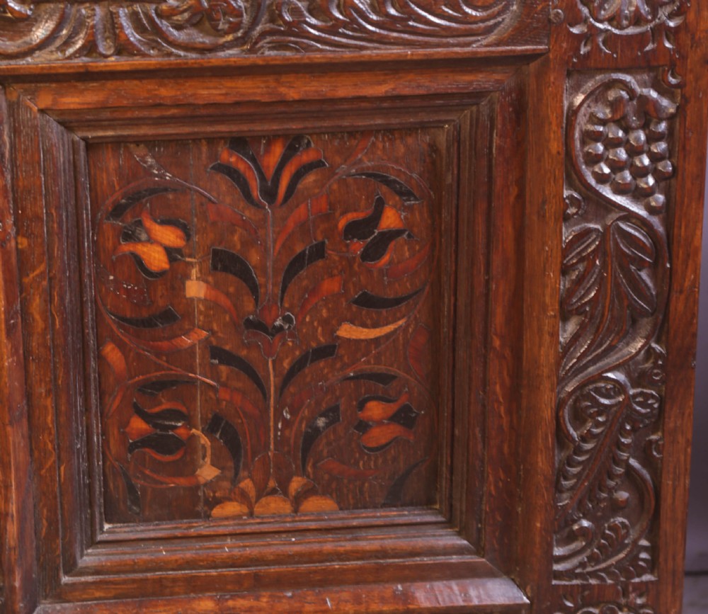 An antique carved oak coffer featuring three floral inlaid panels, carved frieze work, and two lower drawers with brass handles. View X