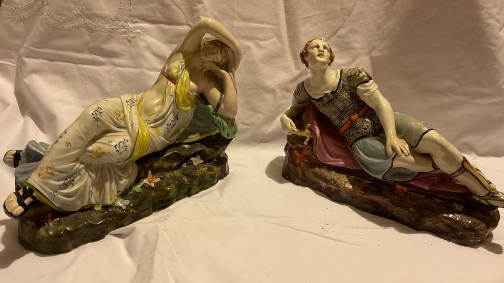 A pair of antique ceramic reclining figures depicting Mark Antony and Cleopatra on moulded bases.