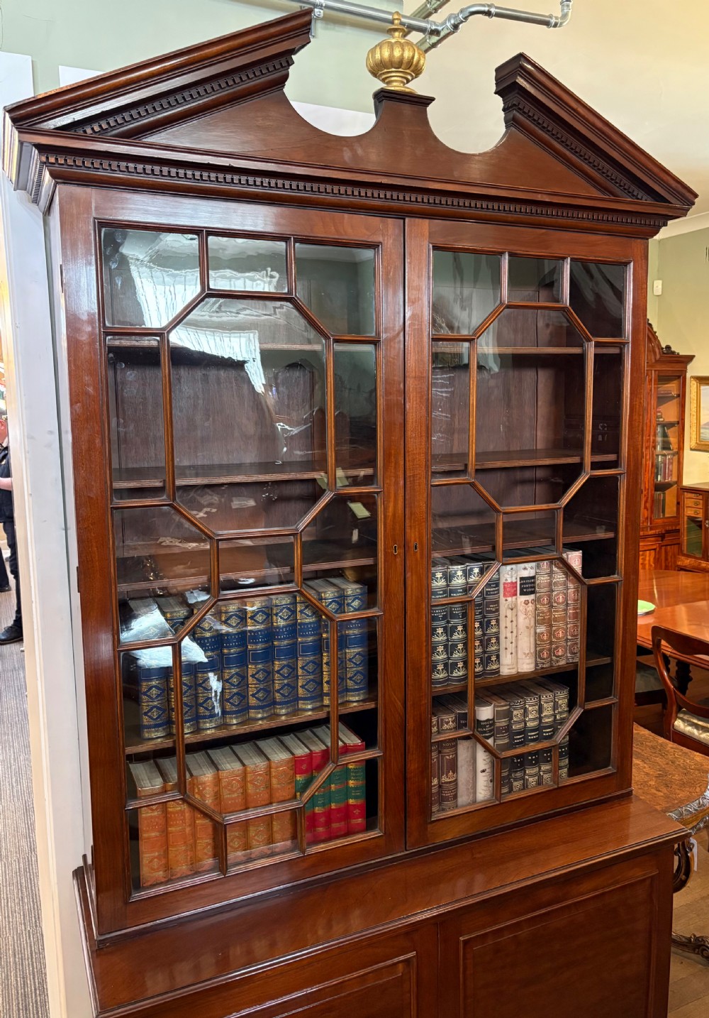 A Georgian mahogany library bookcase featuring a broken swan-neck pediment with a central gilded urn finial, two astragal glazed upper doors, and two solid panelled cabinet doors below. View IV