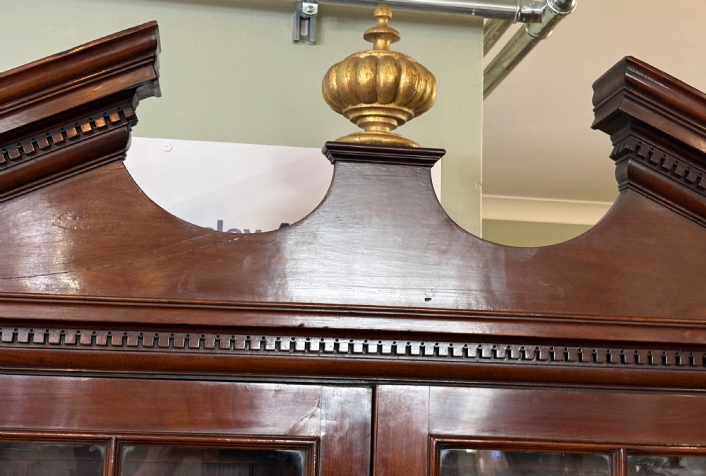 A Georgian mahogany library bookcase featuring a broken swan-neck pediment with a central gilded urn finial, two astragal glazed upper doors, and two solid panelled cabinet doors below. View VIII