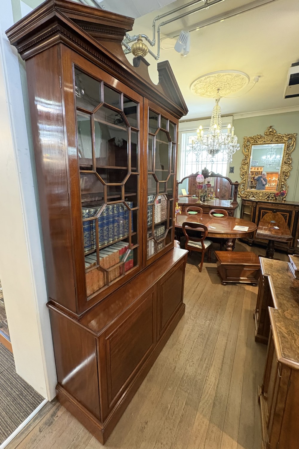 A Georgian mahogany library bookcase featuring a broken swan-neck pediment with a central gilded urn finial, two astragal glazed upper doors, and two solid panelled cabinet doors below. View XII