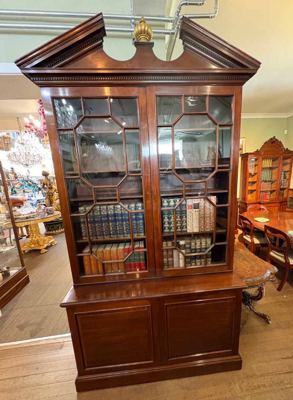 A Georgian mahogany library bookcase featuring a broken swan-neck pediment with a central gilded urn finial, two astragal glazed upper doors, and two solid panelled cabinet doors below. View XIV