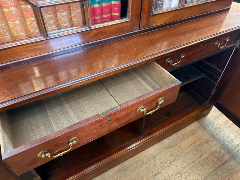 A Georgian mahogany library bookcase featuring a broken swan-neck pediment with a central gilded urn finial, two astragal glazed upper doors, and two solid panelled cabinet doors below. View XV