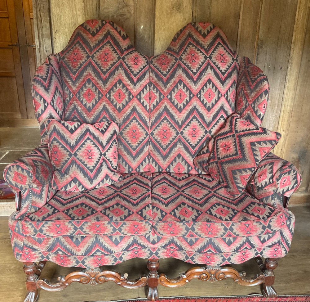 A two-seater high-back wingback sofa featuring bold geometric patterned upholstery in shades of red, black, and cream, set on an ornate carved wood frame with decorative stretchers.