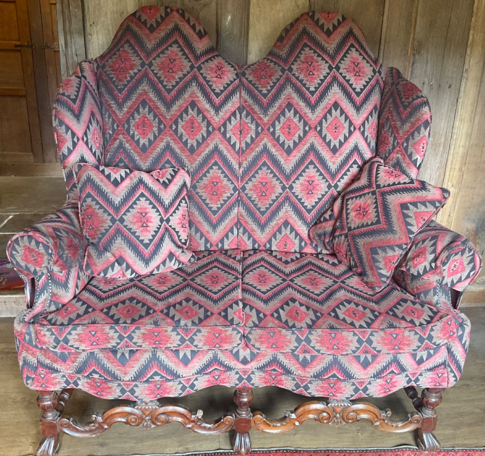A two-seater high-back wingback sofa featuring bold geometric patterned upholstery in shades of red, black, and cream, set on an ornate carved wood frame with decorative stretchers. View II