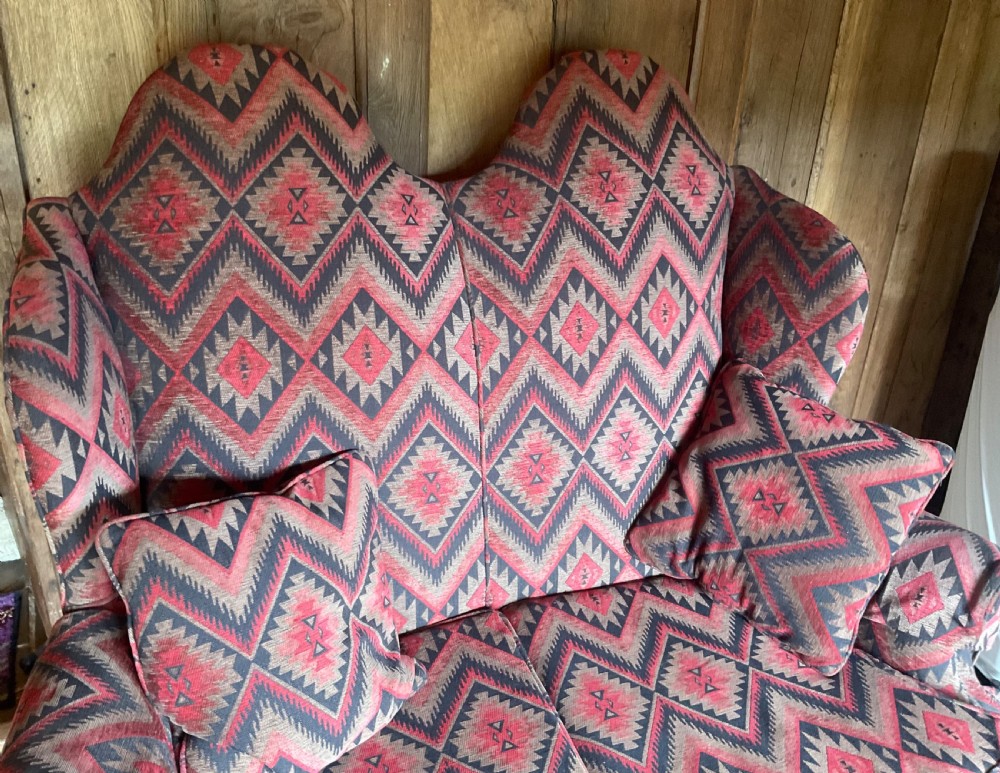 A two-seater high-back wingback sofa featuring bold geometric patterned upholstery in shades of red, black, and cream, set on an ornate carved wood frame with decorative stretchers. View III