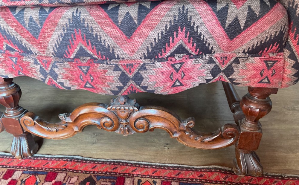 A two-seater high-back wingback sofa featuring bold geometric patterned upholstery in shades of red, black, and cream, set on an ornate carved wood frame with decorative stretchers. View V