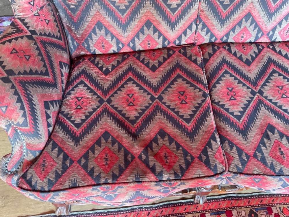 A two-seater high-back wingback sofa featuring bold geometric patterned upholstery in shades of red, black, and cream, set on an ornate carved wood frame with decorative stretchers. View VI