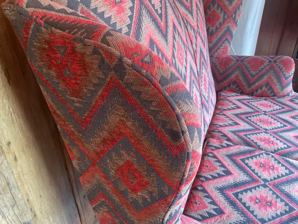 A two-seater high-back wingback sofa featuring bold geometric patterned upholstery in shades of red, black, and cream, set on an ornate carved wood frame with decorative stretchers. View VIII