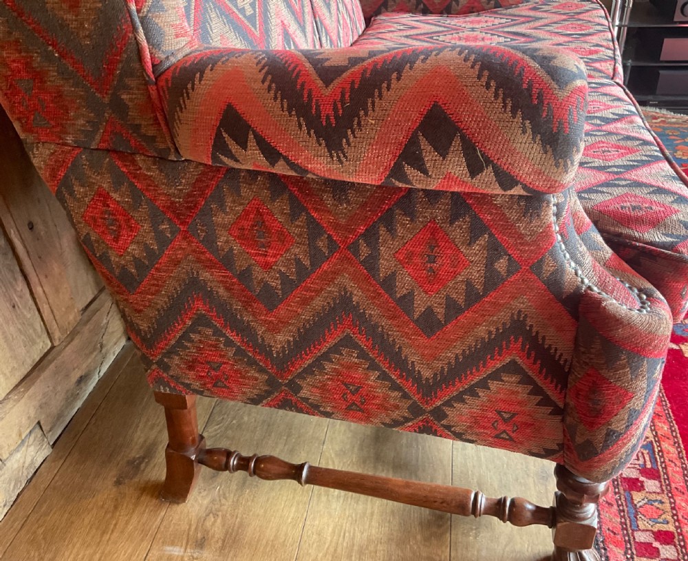 A two-seater high-back wingback sofa featuring bold geometric patterned upholstery in shades of red, black, and cream, set on an ornate carved wood frame with decorative stretchers. View IX