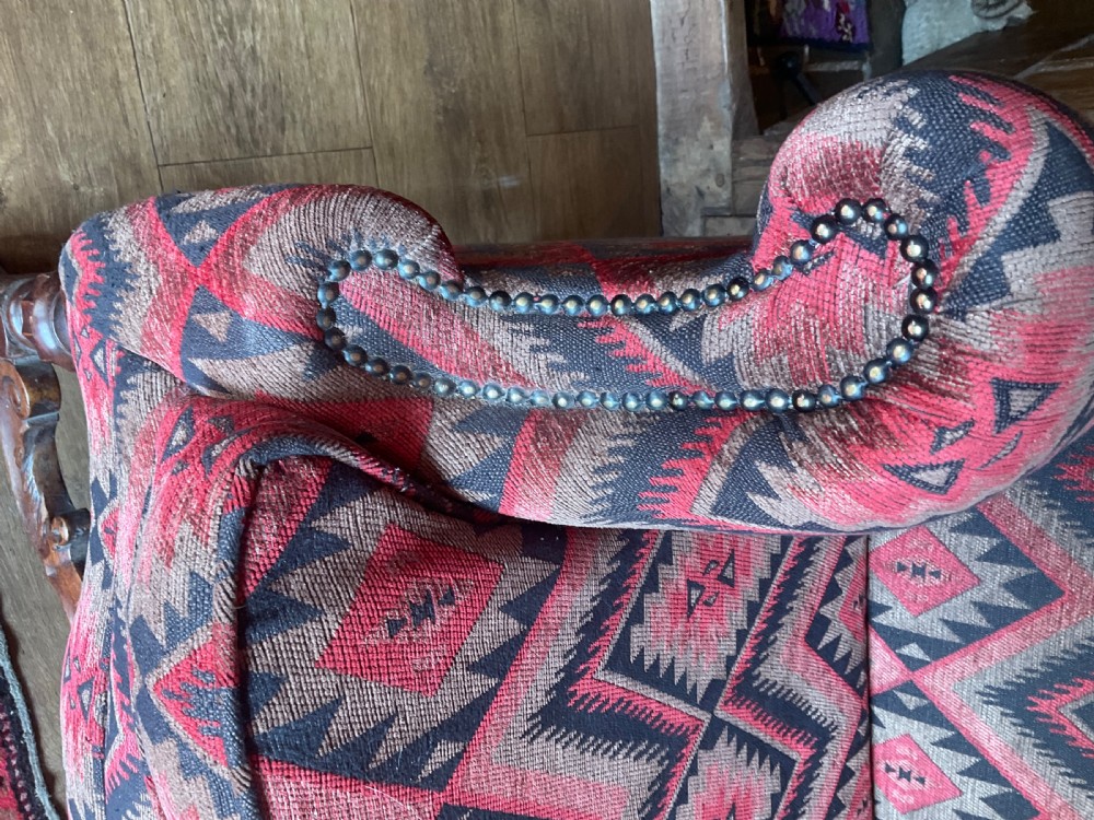 A two-seater high-back wingback sofa featuring bold geometric patterned upholstery in shades of red, black, and cream, set on an ornate carved wood frame with decorative stretchers. View X