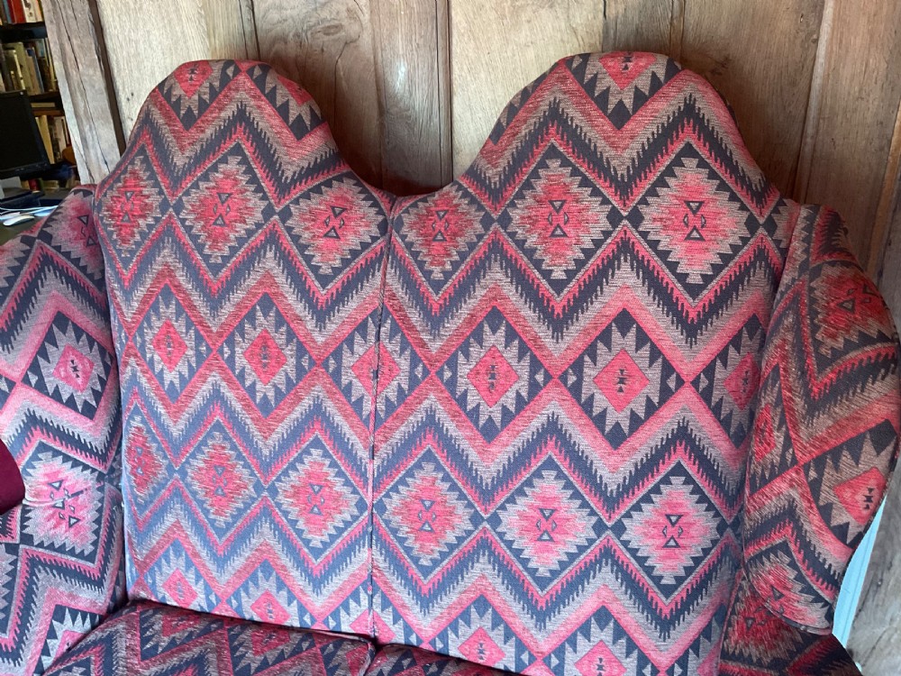 A two-seater high-back wingback sofa featuring bold geometric patterned upholstery in shades of red, black, and cream, set on an ornate carved wood frame with decorative stretchers. View XI