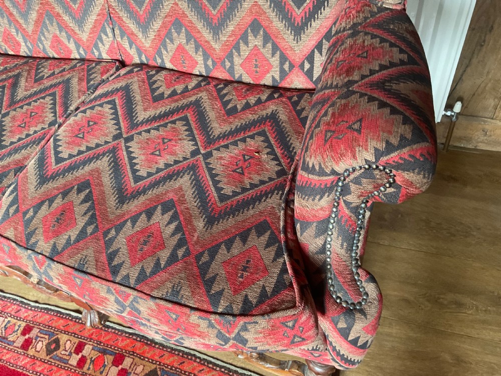 A two-seater high-back wingback sofa featuring bold geometric patterned upholstery in shades of red, black, and cream, set on an ornate carved wood frame with decorative stretchers. View XII