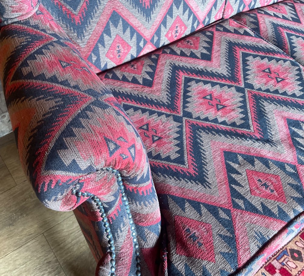 A two-seater high-back wingback sofa featuring bold geometric patterned upholstery in shades of red, black, and cream, set on an ornate carved wood frame with decorative stretchers. View XIII