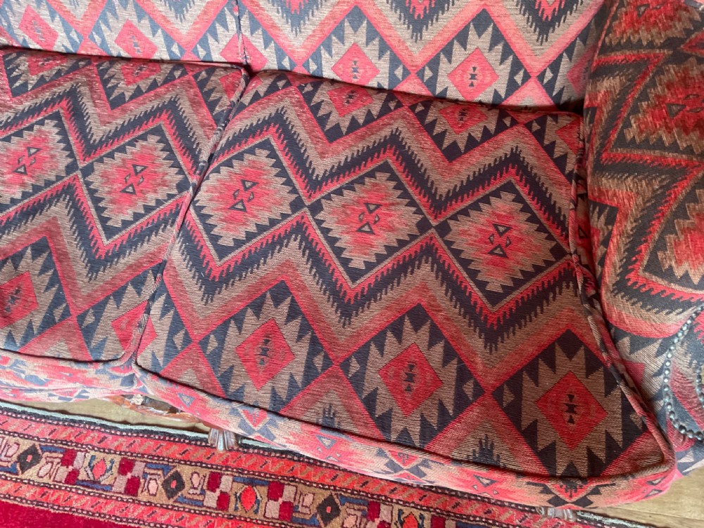 A two-seater high-back wingback sofa featuring bold geometric patterned upholstery in shades of red, black, and cream, set on an ornate carved wood frame with decorative stretchers. View XIV