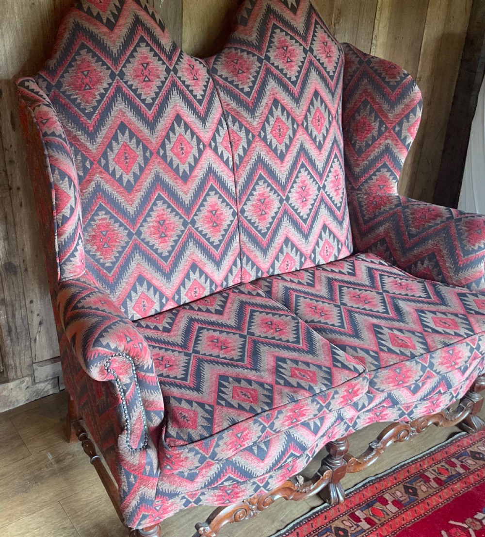 A two-seater high-back wingback sofa featuring bold geometric patterned upholstery in shades of red, black, and cream, set on an ornate carved wood frame with decorative stretchers. View XV