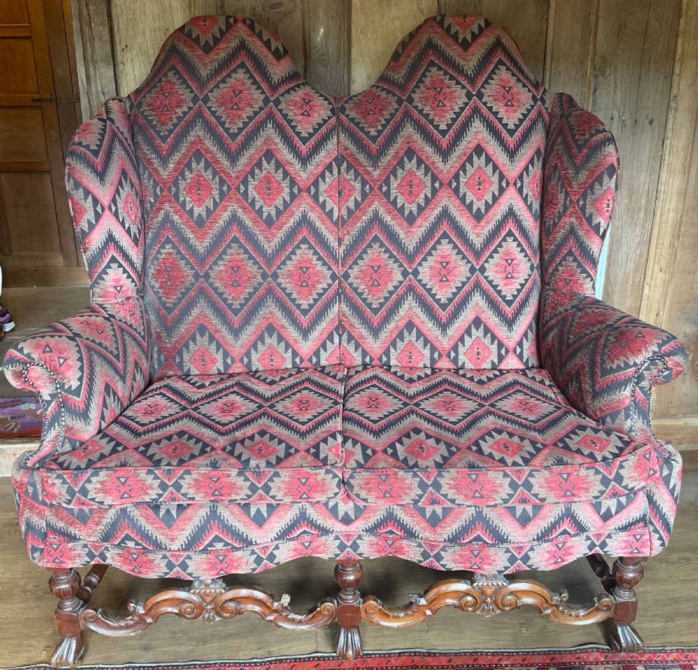 A two-seater high-back wingback sofa featuring bold geometric patterned upholstery in shades of red, black, and cream, set on an ornate carved wood frame with decorative stretchers. View XVI
