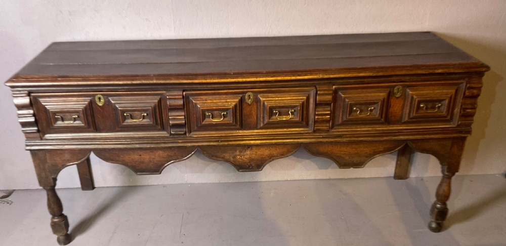 A late seventeenth-century dark oak serving dresser featuring three geometric moulded drawers with brass handles and a scalloped lower apron set on turned legs.