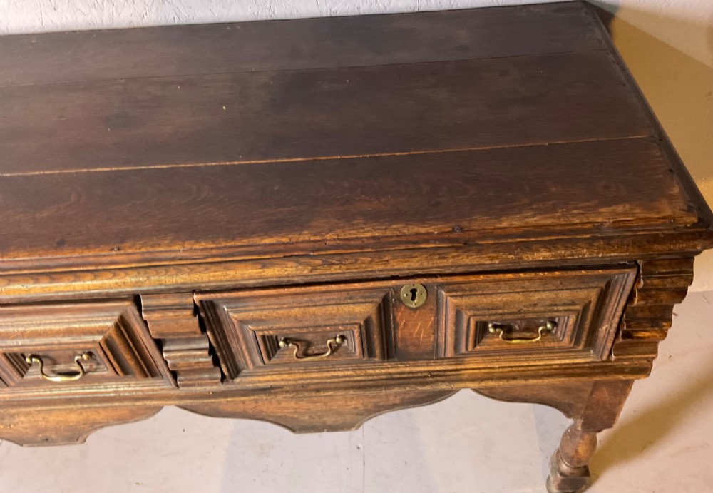 A late seventeenth-century dark oak serving dresser featuring three geometric moulded drawers with brass handles and a scalloped lower apron set on turned legs. View II