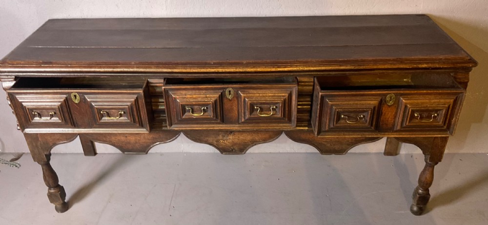 A late seventeenth-century dark oak serving dresser featuring three geometric moulded drawers with brass handles and a scalloped lower apron set on turned legs. View III