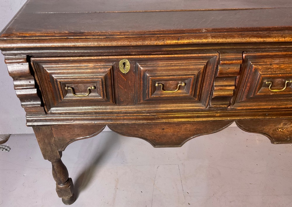 A late seventeenth-century dark oak serving dresser featuring three geometric moulded drawers with brass handles and a scalloped lower apron set on turned legs. View V