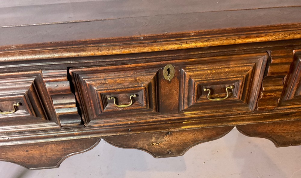 A late seventeenth-century dark oak serving dresser featuring three geometric moulded drawers with brass handles and a scalloped lower apron set on turned legs. View VI