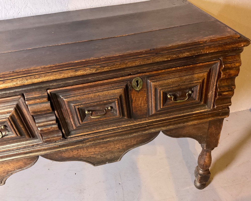 A late seventeenth-century dark oak serving dresser featuring three geometric moulded drawers with brass handles and a scalloped lower apron set on turned legs. View VII