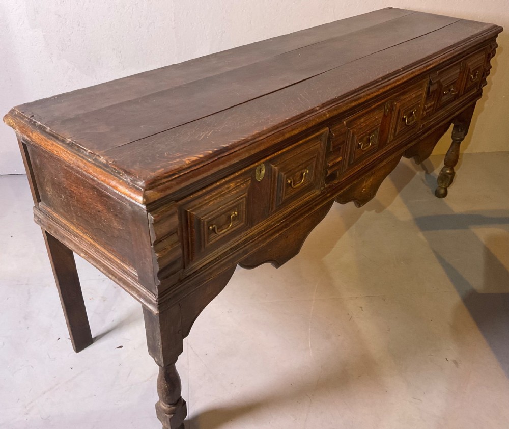 A late seventeenth-century dark oak serving dresser featuring three geometric moulded drawers with brass handles and a scalloped lower apron set on turned legs. View IX