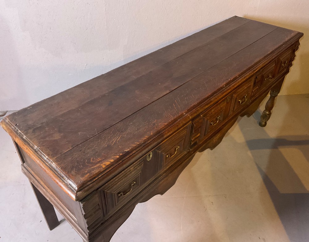 A late seventeenth-century dark oak serving dresser featuring three geometric moulded drawers with brass handles and a scalloped lower apron set on turned legs. View X
