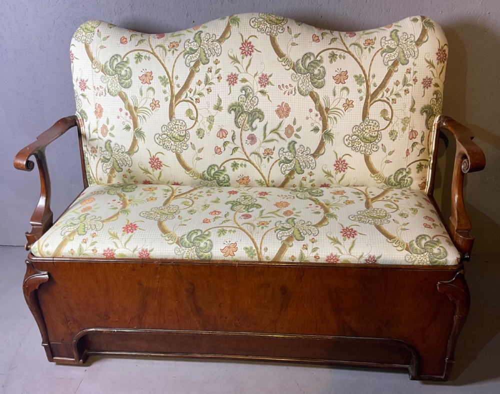 A Queen Anne style walnut settee featuring contoured wooden arms, a shaped upholstered backrest, and botanical floral pattern fabric.