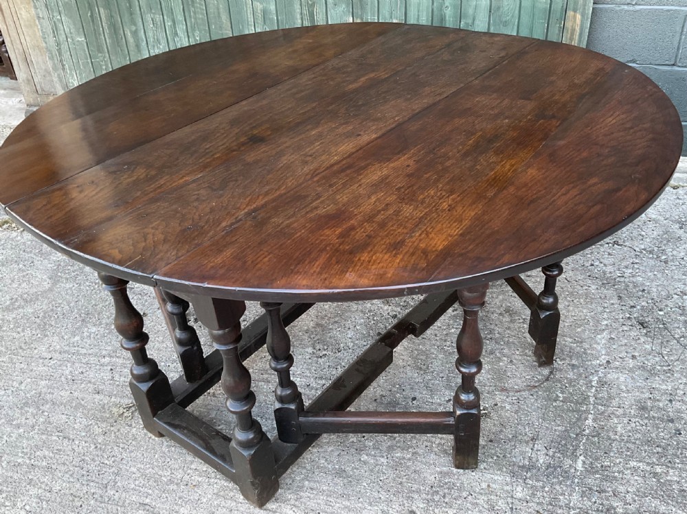 Underside view of a dark brown oak gateleg table showing baluster turned legs, stretchers, hand-forged iron hinges, and hinged drop leaves. View III