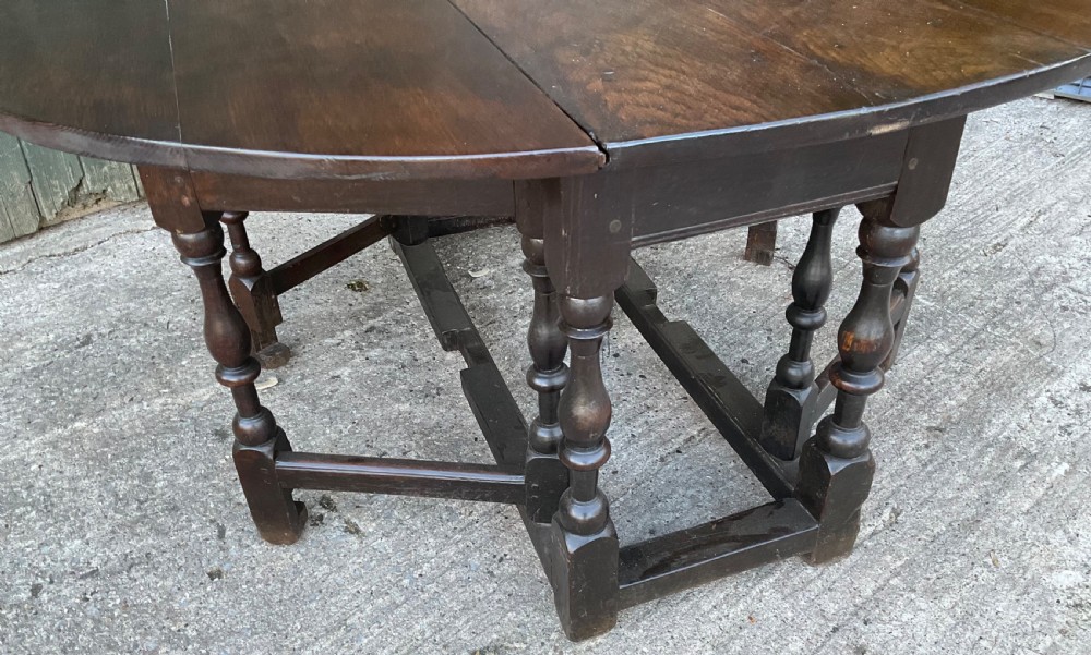 Underside view of a dark brown oak gateleg table showing baluster turned legs, stretchers, hand-forged iron hinges, and hinged drop leaves. View VII