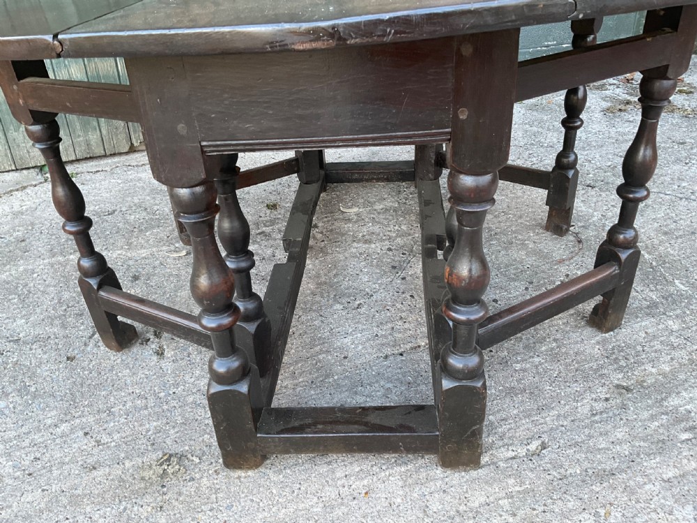 Underside view of a dark brown oak gateleg table showing baluster turned legs, stretchers, hand-forged iron hinges, and hinged drop leaves. View X