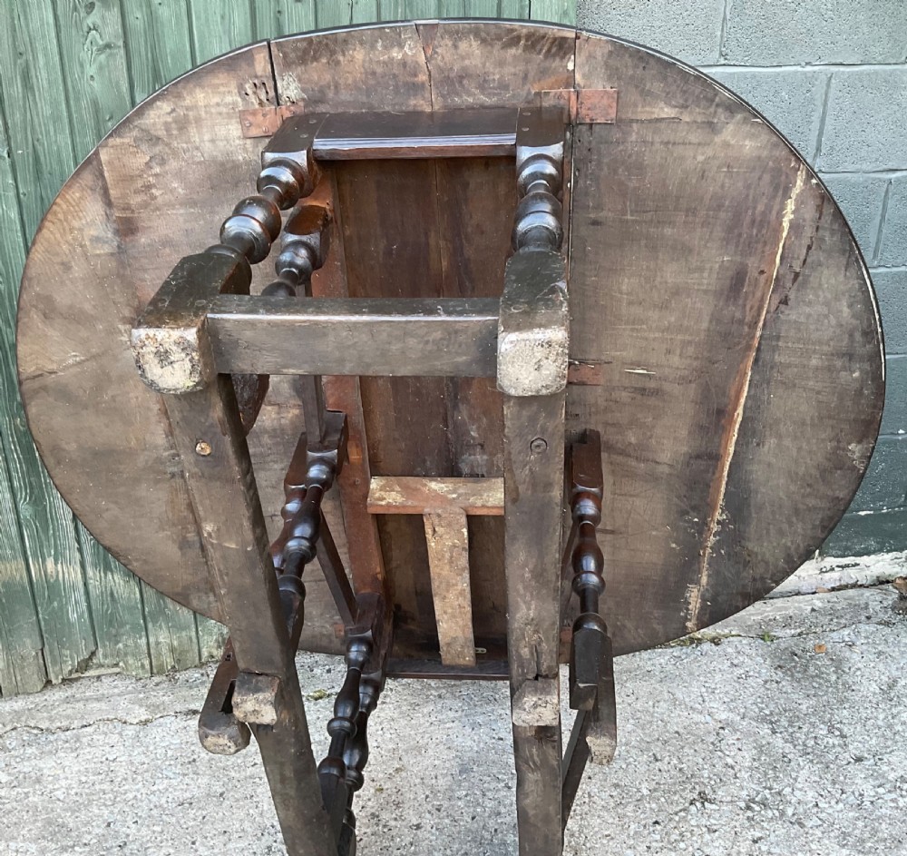 Underside view of a dark brown oak gateleg table showing baluster turned legs, stretchers, hand-forged iron hinges, and hinged drop leaves.