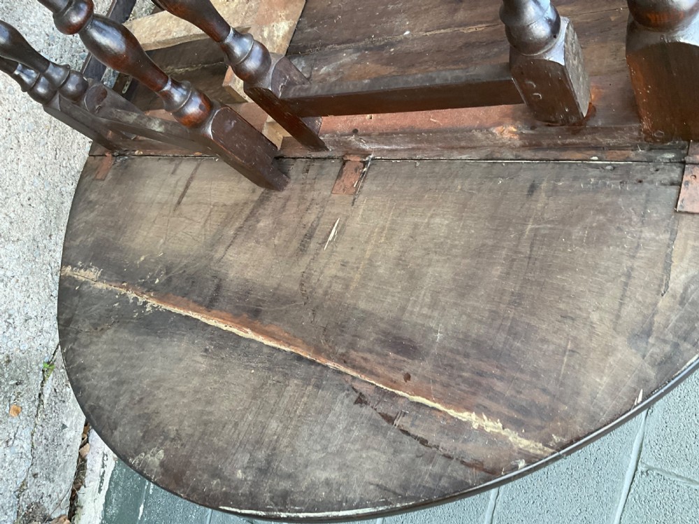 Underside view of a dark brown oak gateleg table showing baluster turned legs, stretchers, hand-forged iron hinges, and hinged drop leaves. View XVI