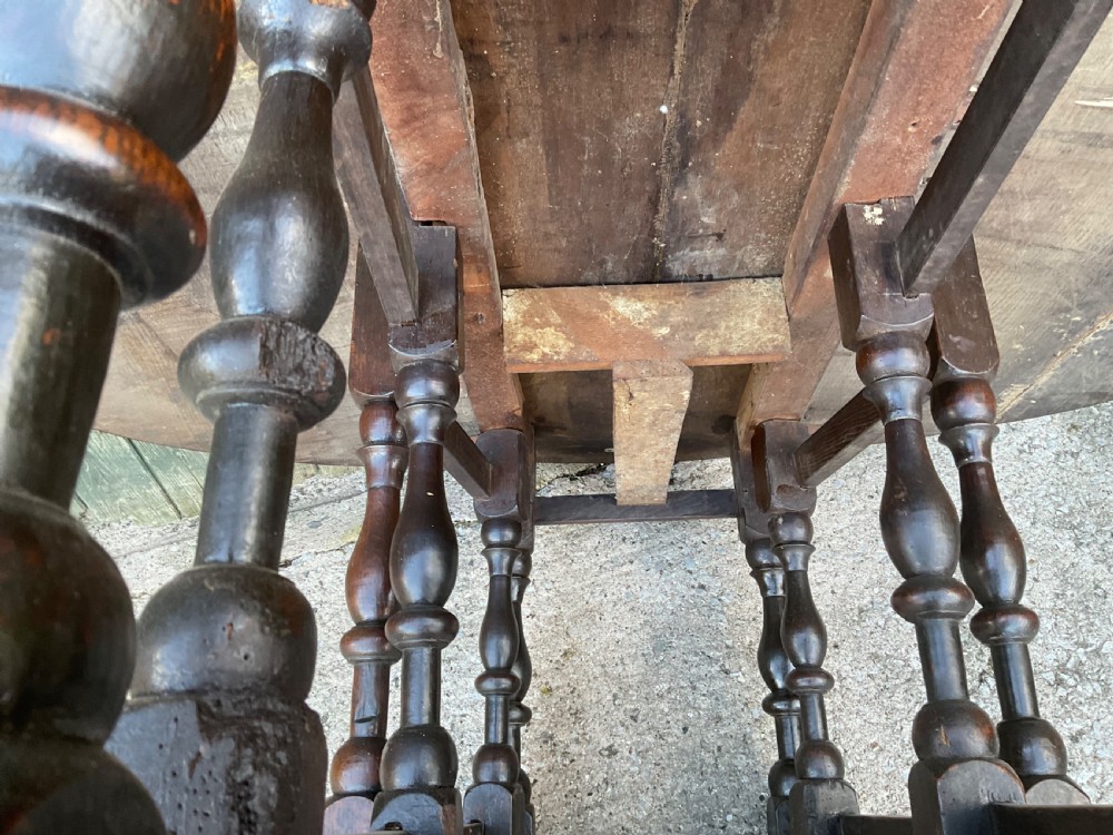 Underside view of a dark brown oak gateleg table showing baluster turned legs, stretchers, hand-forged iron hinges, and hinged drop leaves. View XVII