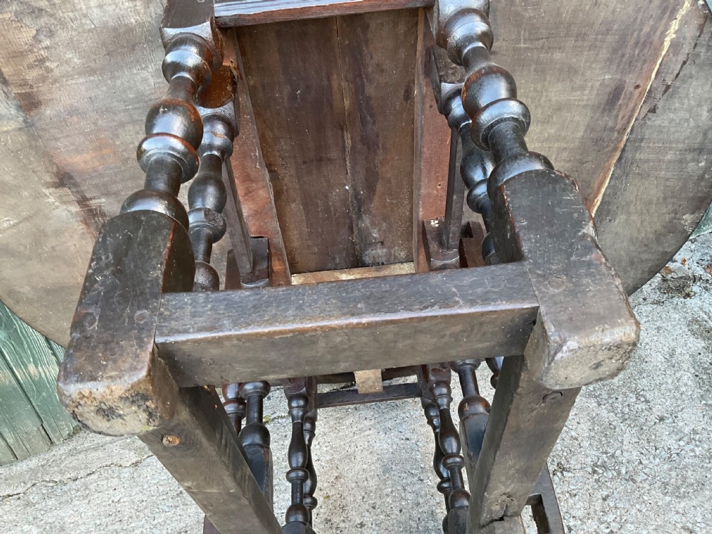 Underside view of a dark brown oak gateleg table showing baluster turned legs, stretchers, hand-forged iron hinges, and hinged drop leaves. View XXI