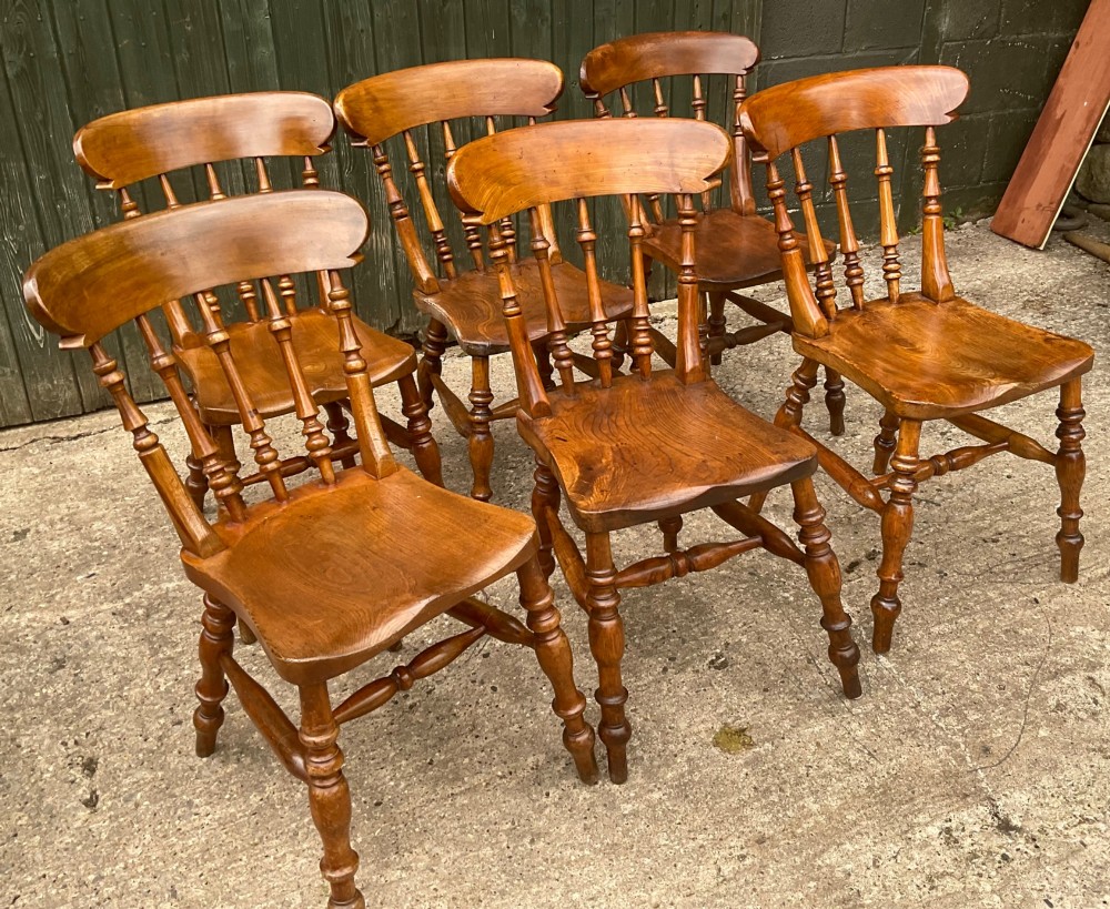A set of six warm brown wooden dining chairs featuring spindle backrests, contoured wooden seats, and turned legs. View V
