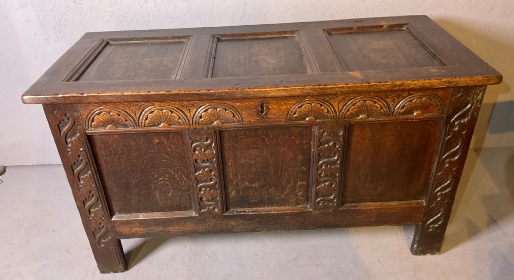 A dark carved 17th-century oak coffer featuring three panelled sections, a hinged lid, and decorative lunette carving along the frieze.