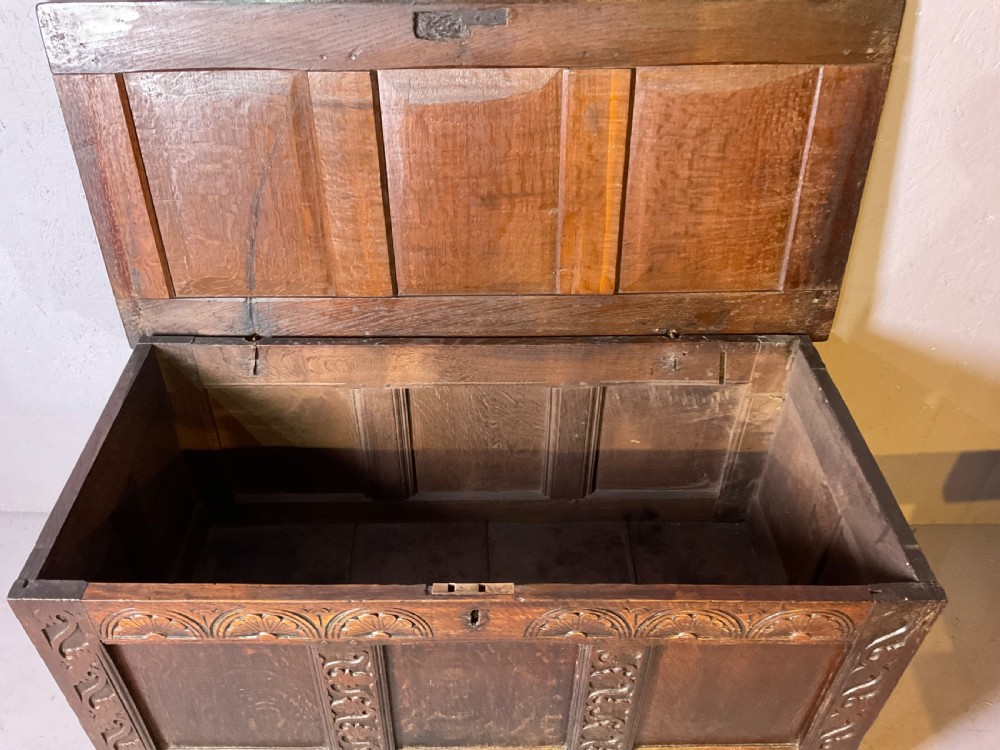 A dark carved 17th-century oak coffer featuring three panelled sections, a hinged lid, and decorative lunette carving along the frieze. View V