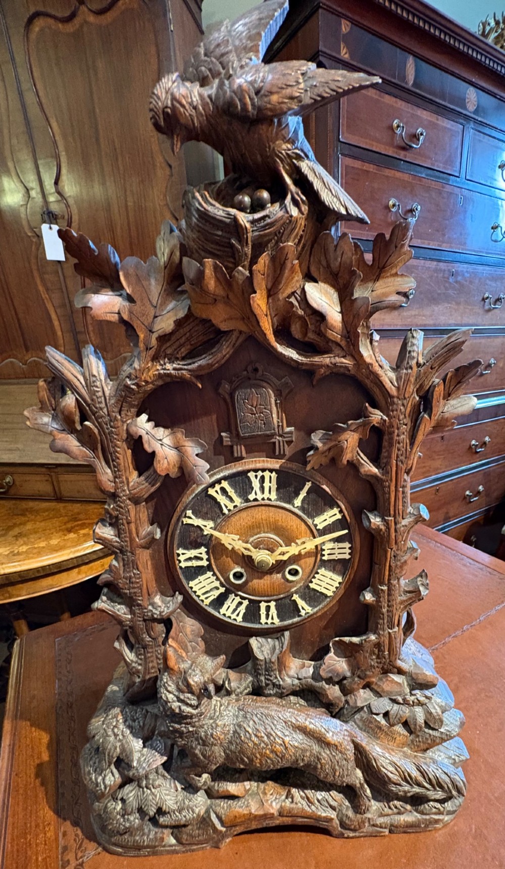 An intricately hand-carved dark wood Black Forest cuckoo clock featuring a bird atop a nest, a framed dial with Roman numerals, and a fox figure at the base.