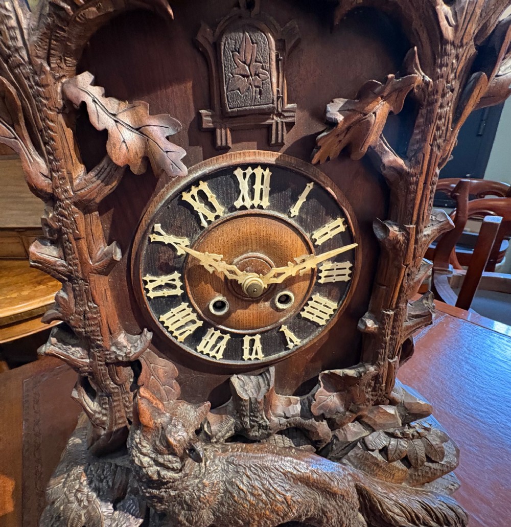 An intricately hand-carved dark wood Black Forest cuckoo clock featuring a bird atop a nest, a framed dial with Roman numerals, and a fox figure at the base. View III
