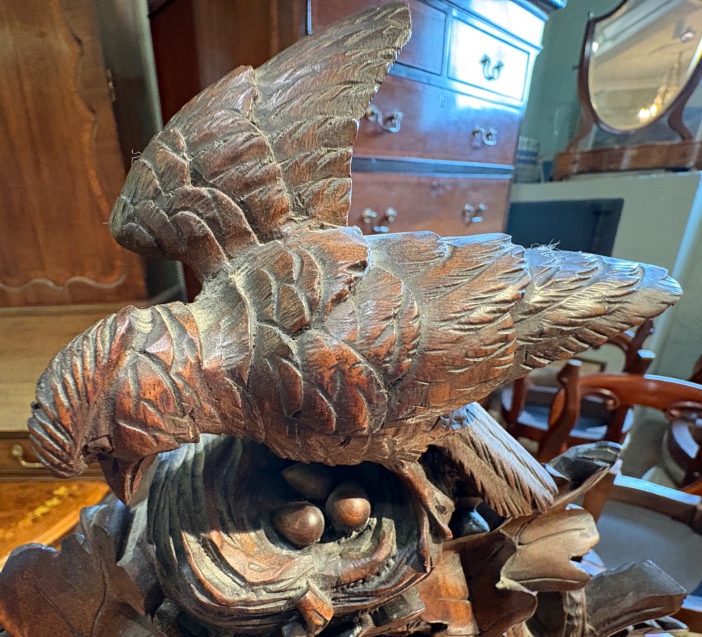 An intricately hand-carved dark wood Black Forest cuckoo clock featuring a bird atop a nest, a framed dial with Roman numerals, and a fox figure at the base. View V