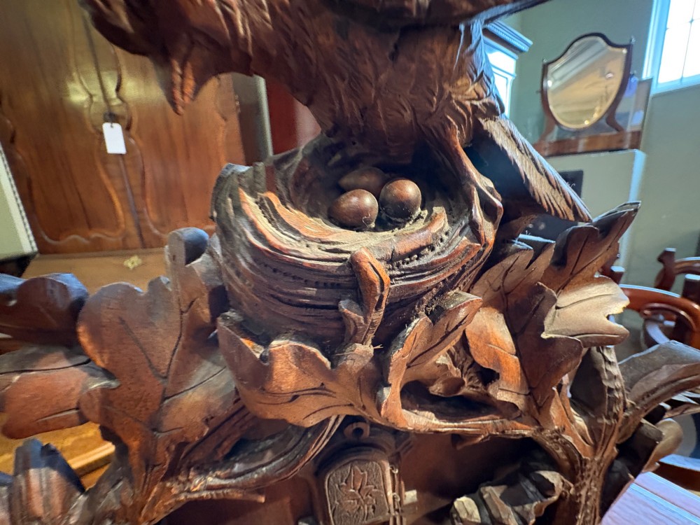 An intricately hand-carved dark wood Black Forest cuckoo clock featuring a bird atop a nest, a framed dial with Roman numerals, and a fox figure at the base. View VI