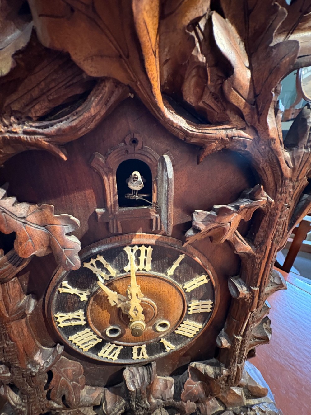 An intricately hand-carved dark wood Black Forest cuckoo clock featuring a bird atop a nest, a framed dial with Roman numerals, and a fox figure at the base. View VII